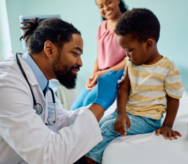 Doctor administering a vaccine to a child on an exam table while an adult looks on in a clinic.