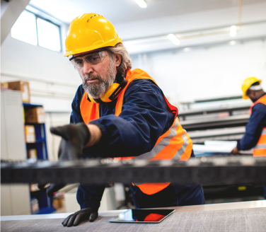 Worker in a hard hat and high-visibility vest handling materials on a factory floor with a tablet nearby