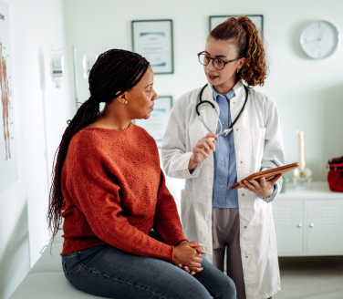 Healthcare provider in a white coat reviewing a clipboard with a person seated in a clinical exam room