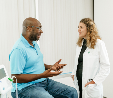 Marathon Health doctor in a white coat speaking with a seated individual in a clinical exam room with medical equipment nearby.