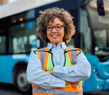 Worker in a high-visibility safety vest standing with arms crossed in front of a transit bus, smiling at the camera