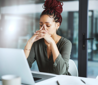 Tired employee sitting at a desk with a laptop, hands raised to face, in an office setting.