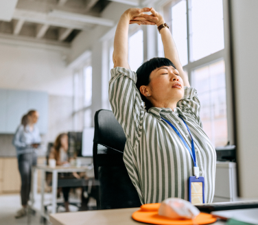 Employee stretching arms overhead at a workplace desk, with coworkers and bright windows visible in the background.