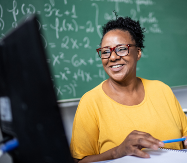 Teacher smiling at a computer screen while writing notes, seated in front of a chalkboard covered in math equations.