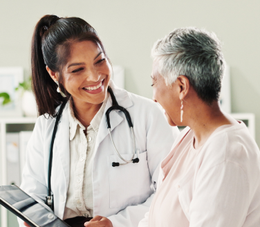 Healthcare provider in a white coat and stethoscope reviewing a tablet with a person in a clinic setting