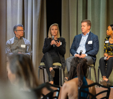 Four panelists seated on a stage speak with microphones before an audience