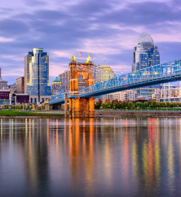 A suspension bridge with lit brick towers spans a calm river at dusk, with a city skyline glowing against a purple sky.