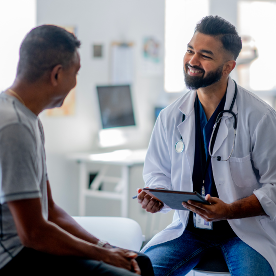 A smiling healthcare provider in a white coat and stethoscope holds a tablet while speaking with an ADVICS employee in a bright clinic.