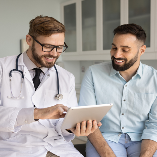 A healthcare provider in a white coat and stethoscope reviews a tablet with a smiling person in a clinical setting