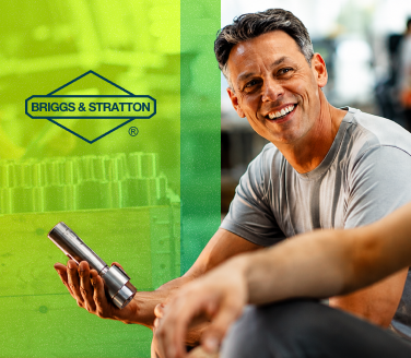 Smiling person holding a metal part in a gym setting, with Briggs & Stratton branding visible