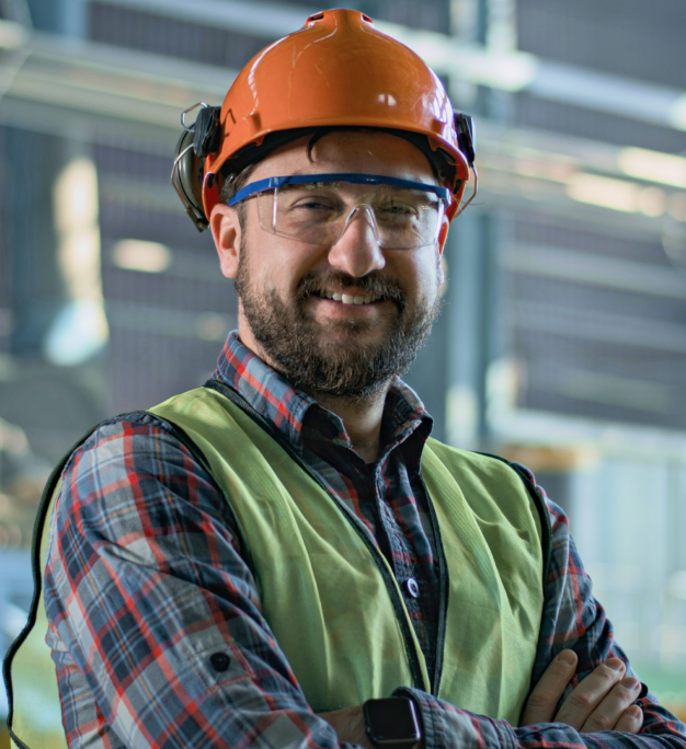 Smiling person in orange hard hat, safety glasses, and hi-vis vest stands arms crossed in a Briggs & Stratton facility.