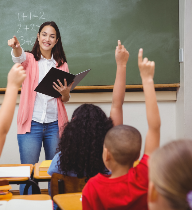 Smiling teacher holding a binder points to students raising hands in a classroom with math equations on the chalkboard