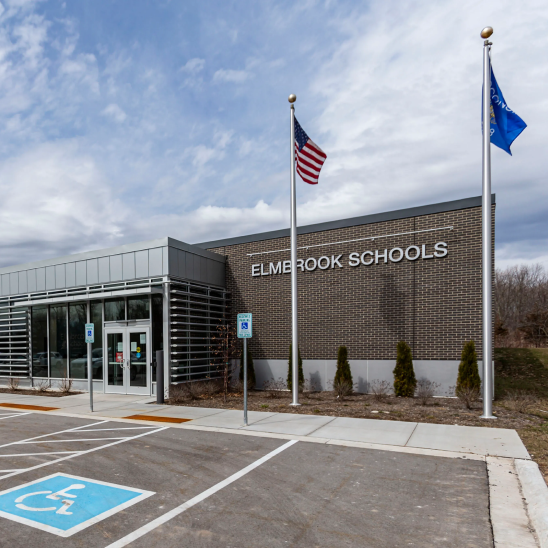 Elmbrook Schools brick building exterior with signage, flagpoles flying the US and state flags, and accessible parking spaces