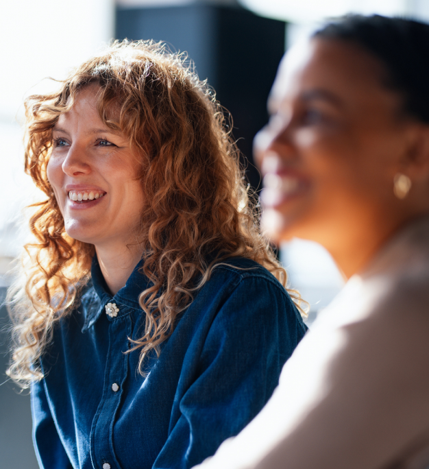 Two people smiling in a bright indoor setting, one in focus wearing a denim shirt with curly auburn hair