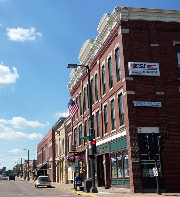 Tree-lined small-town main street with historic red-brick buildings, an American flag, and a traffic light
