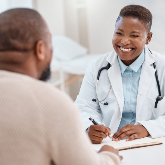 Smiling healthcare provider in white coat and stethoscope writes notes while speaking with a person across a desk