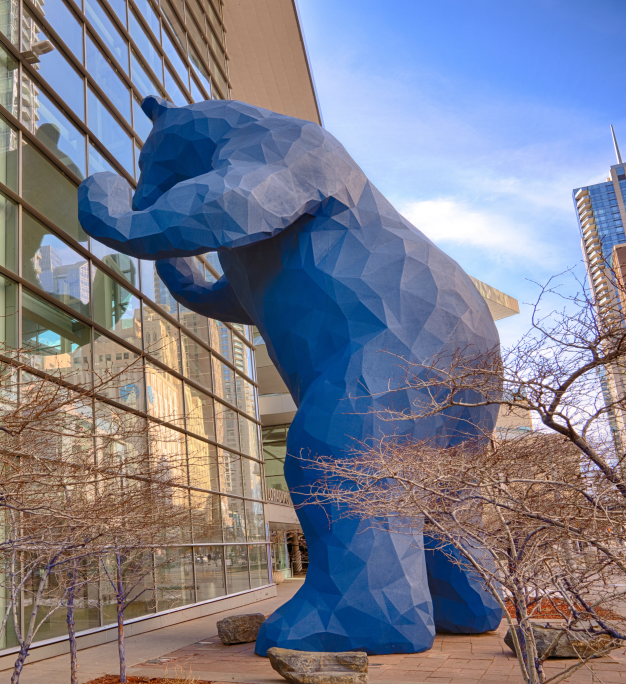 Large blue geometric bear sculpture peering into a glass building facade surrounded by bare trees and city skyline