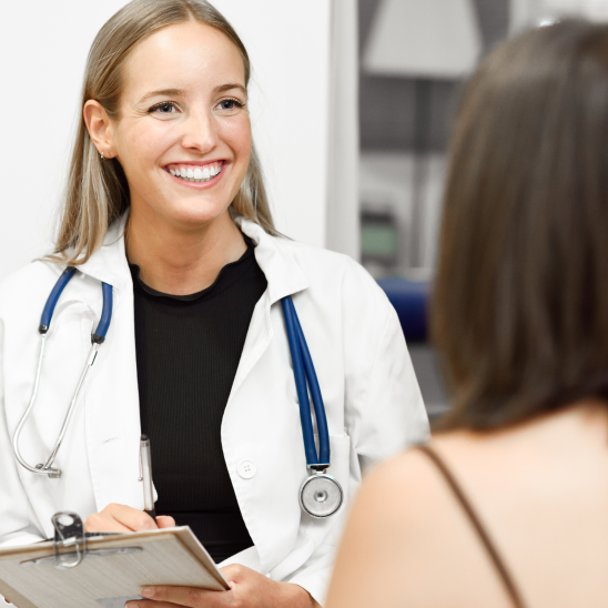 Smiling healthcare provider in white coat and stethoscope holds clipboard while speaking with a person in a clinical setting