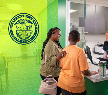 Two people at a reception desk inside a Mecklenburg County facility, with the county seal visible on a green-tinted wall