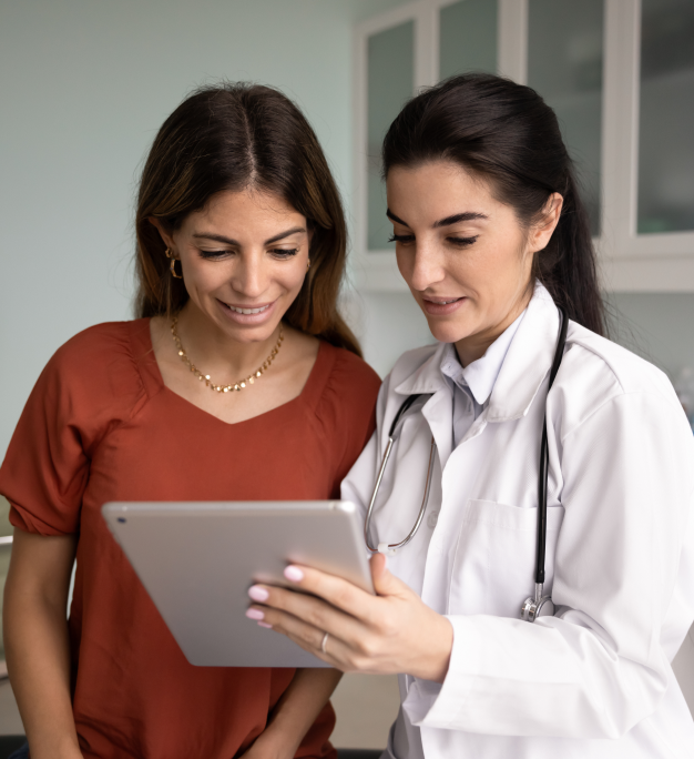 A healthcare provider in a white coat and stethoscope shares a tablet screen with a smiling person in a clinical setting