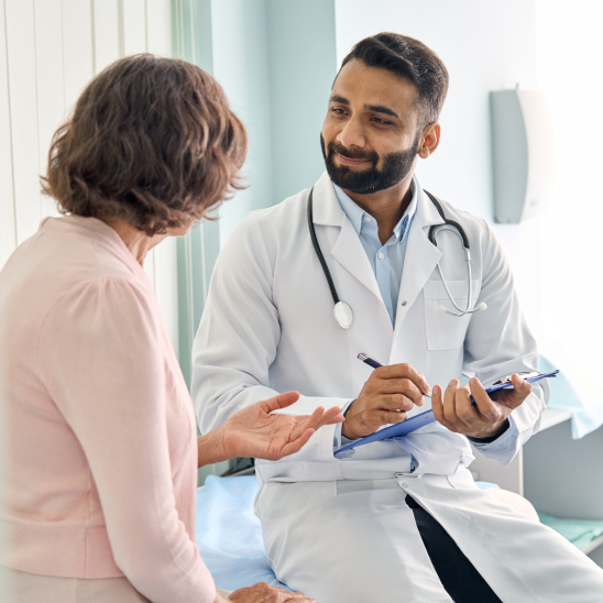 A healthcare provider in a white coat and stethoscope holds a clipboard while speaking with a person in a clinical setting