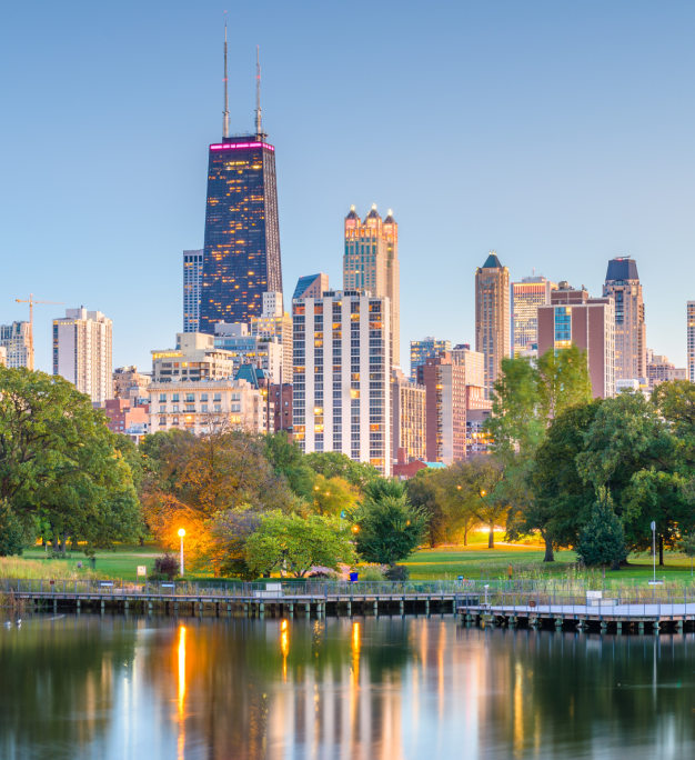 City skyline with tall skyscrapers rising behind a green park, wooden dock, and calm reflecting water at dusk