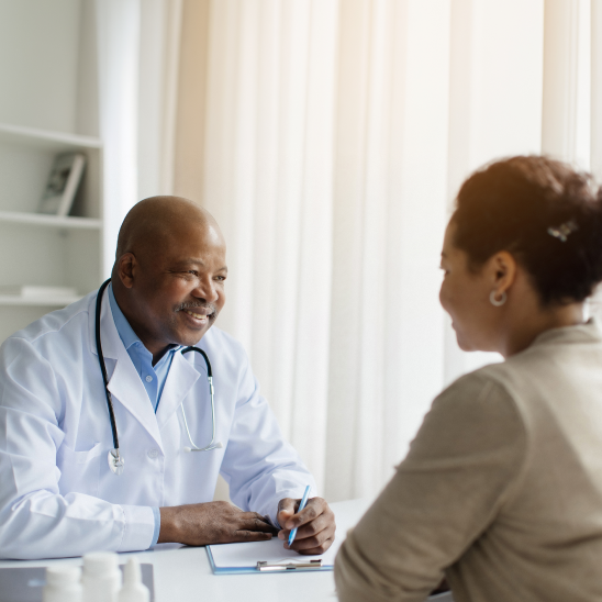 A smiling healthcare provider in a white coat and stethoscope takes notes while speaking with a person seated across a desk