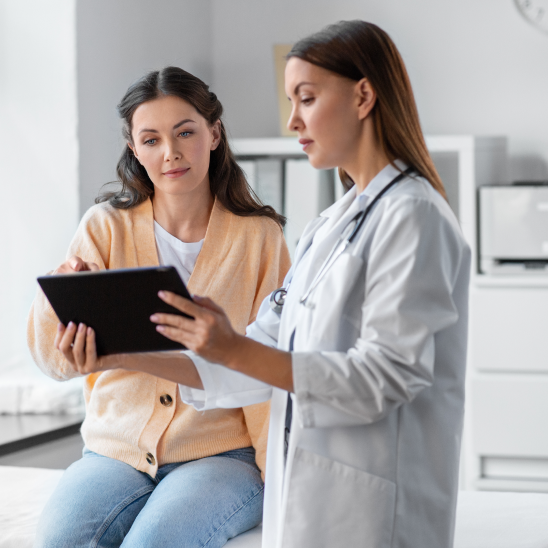 A healthcare provider in a white coat with stethoscope reviews a tablet with a person seated in a bright clinical room
