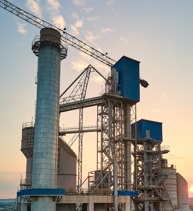 Large industrial facility with tall silos, steel framework, blue structures, and a crane against a sunset sky