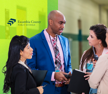 Three professionals in conversation near an Escambia County Public Schools branded green display panel, each holding folders