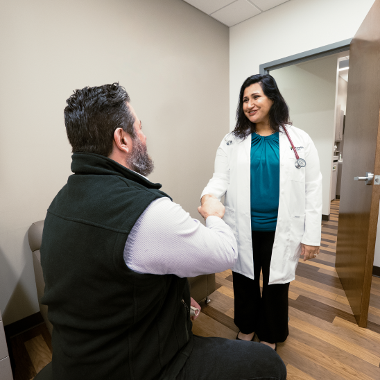 A seated person shakes hands with a smiling healthcare provider in a white coat and stethoscope inside a clinic room