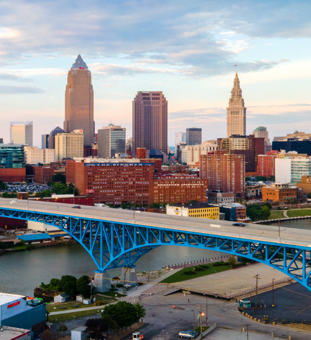 Aerial view of a city skyline at dusk featuring a blue steel arch bridge spanning a calm river