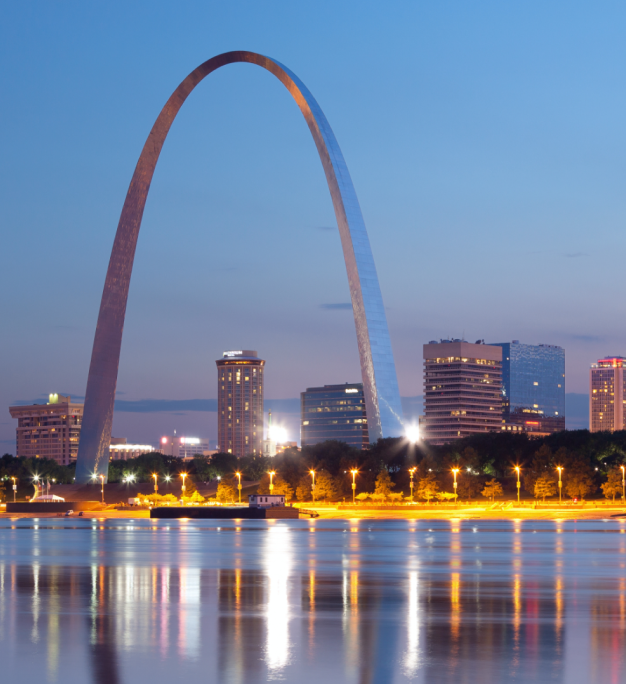 The Gateway Arch rises over the St. Louis skyline at dusk, with city lights reflecting on the river below