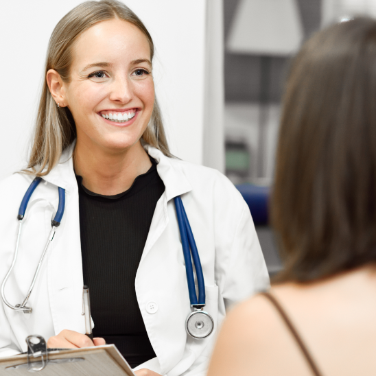 A smiling healthcare provider in a white coat and stethoscope holds a clipboard while speaking with another person