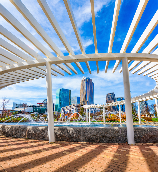 White pergola frames a city park fountain with a downtown skyline and blue sky in the background