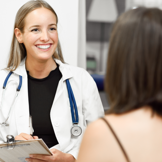Smiling healthcare provider in white coat and stethoscope holds clipboard while speaking with a person seated nearby