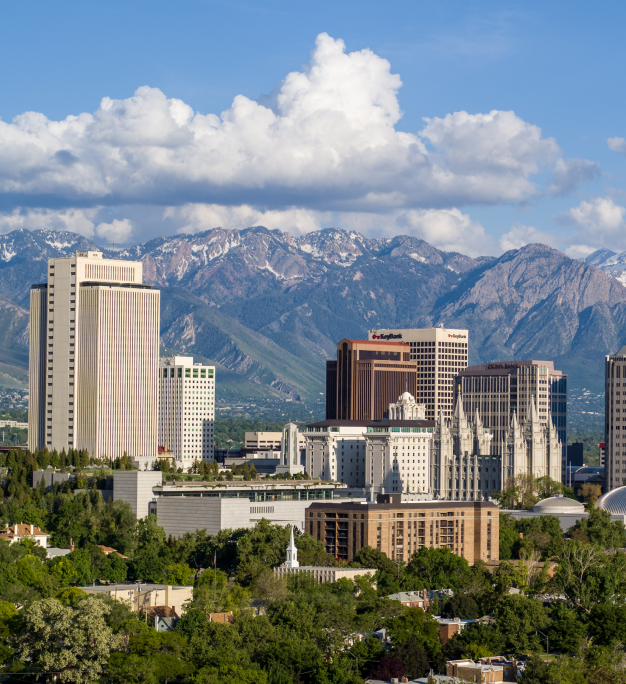 City skyline with tall buildings and tree-lined streets set against snow-capped mountain peaks under a partly cloudy sky.