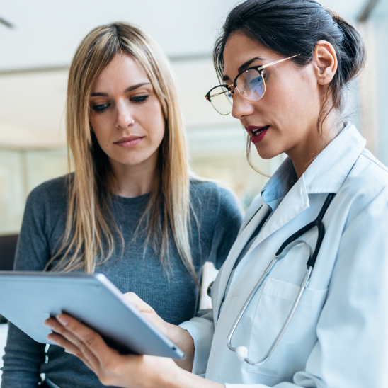 A CHG Healthcare provider in a white coat and stethoscope reviews a tablet with a person in a clinic setting