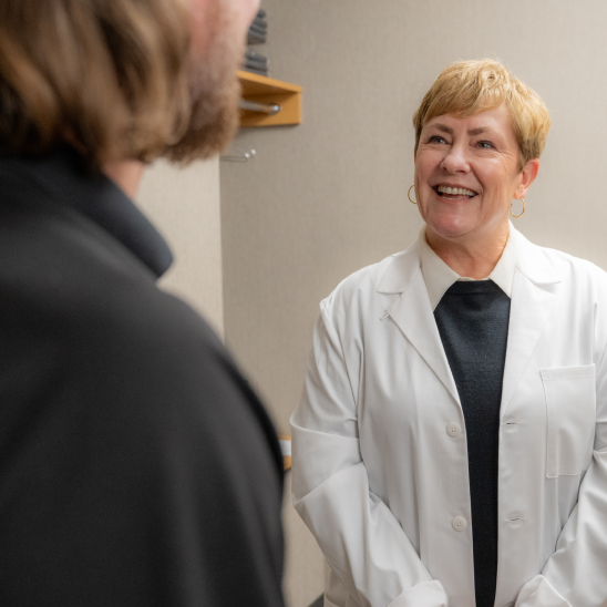 A smiling healthcare provider in a white coat speaks with a person in a dark jacket inside a clinical room