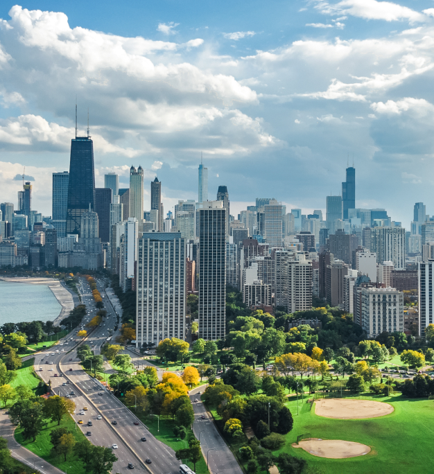 Aerial view of a city skyline with a lakefront parkway, green parks, and a partly cloudy sky