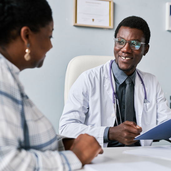 A smiling healthcare provider in a white coat and stethoscope reviews a clipboard while speaking with a person across a desk