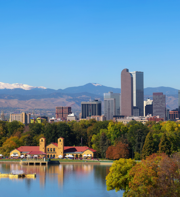 City skyline with tall buildings, a lakeside pavilion, autumn trees, and snow-capped mountains under a clear blue sky