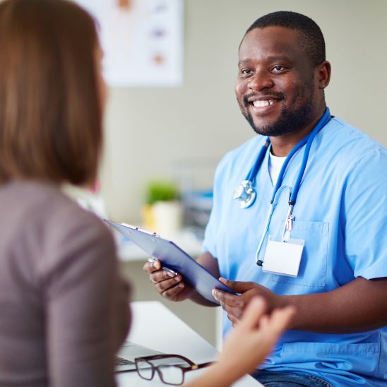 A smiling healthcare provider in blue scrubs holds a clipboard while speaking with a person