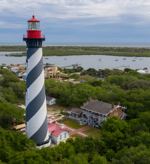 Aerial view of a black-and-white spiral lighthouse with a red top near a coastal waterway and lush trees