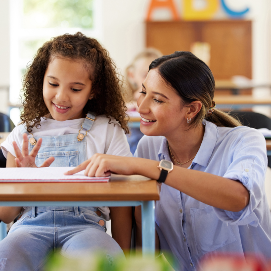 A professional and a student smile together over an open book at a classroom desk
