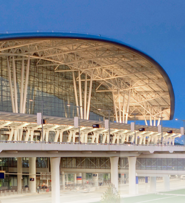 Modern terminal building with a sweeping curved roof and white structural columns lit at dusk against a deep blue sky