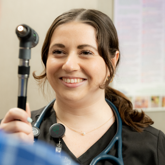 Smiling healthcare provider in black scrubs holding an otoscope with a stethoscope around their neck