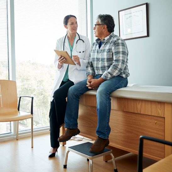 Provider holding a tablet talks with a person seated on an exam table beside a bright window