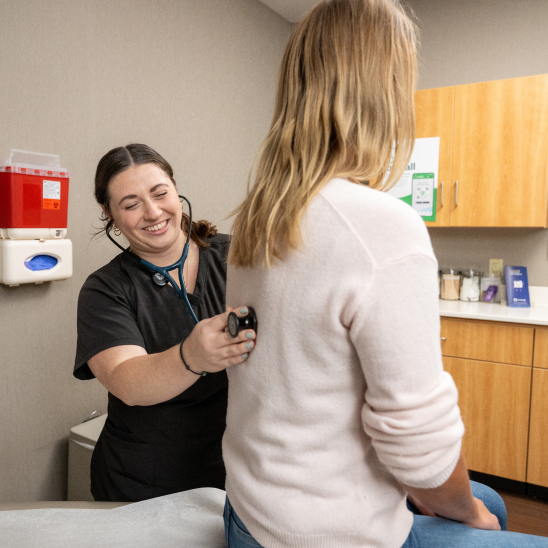 Healthcare provider listens with a stethoscope to the back of a person seated in an exam room