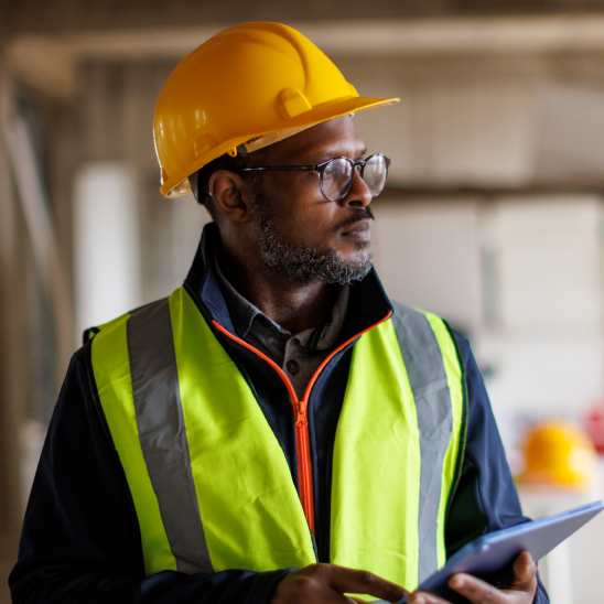 A worker in a yellow hard hat, high-visibility vest, and glasses holds a tablet indoors